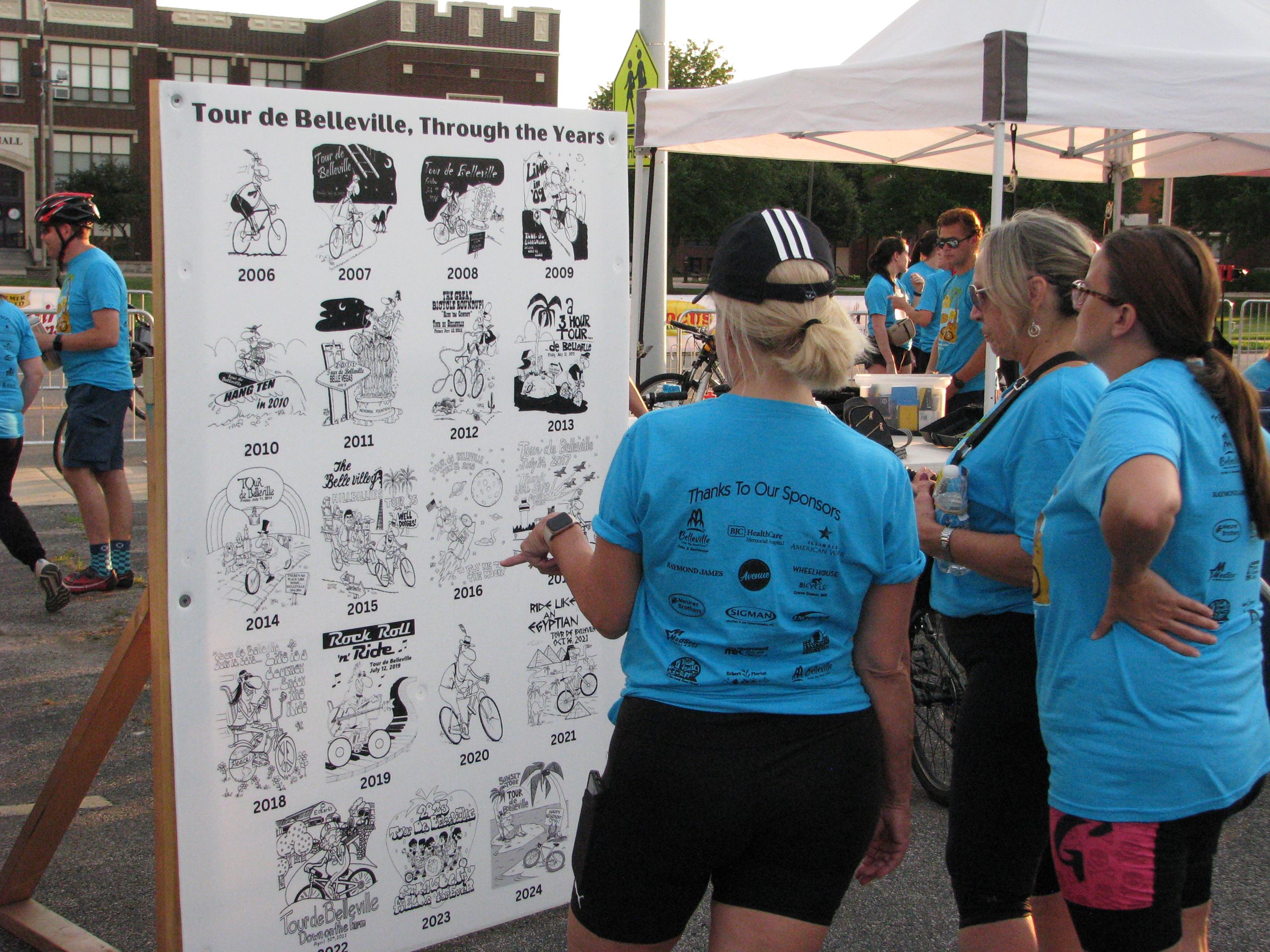 Three women in blue Tour de Belleville t-shirts looking at past event photos
